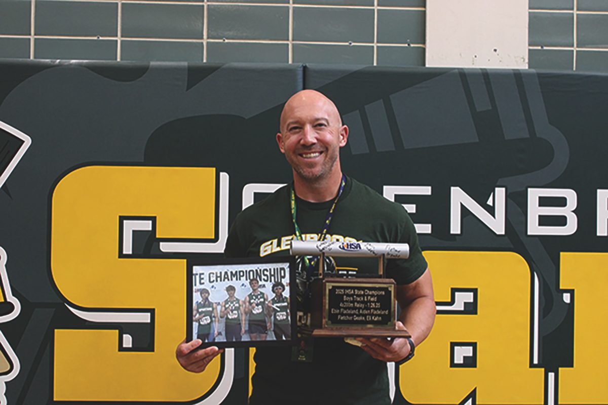 Sean Brandt, head coach of boys track and field, holds the IHSA Boys Track and Field State Championship trophy alongside a photo of the 4x200 relay team. The baton placed on top of the trophy is the same one the team used during the race on May 31.